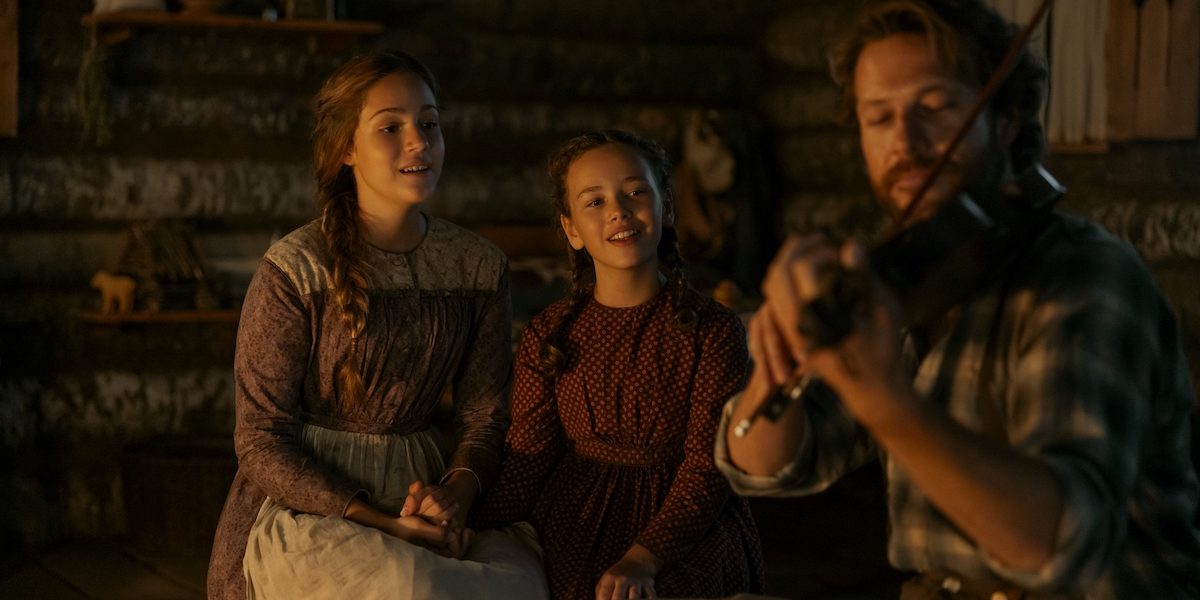 "A man plays the violin for a woman and a young girl inside a rustic wooden cabin, creating a warm, cozy atmosphere with vintage clothing and soft lighting."